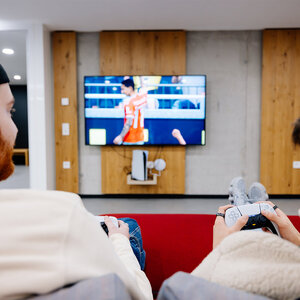 Abbildung zweier Bewohner, die auf einem roten Sitzmöbel sitzen und Videospiele spielen